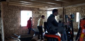 plusieurs personnes avec des brouettes et un homme sur une mini pelleteuse à l'intérieur d'une vieille maison