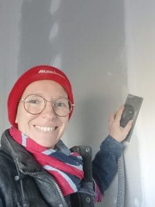 Femme souriante portant des lunettes et un bonnet rouge en train de poncer les bandes de placoplâtres