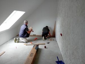Deux hommes à genoux posent du parquet couleur bois, un grand velux éclaire la pièce