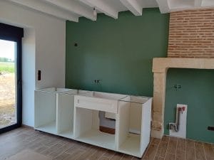Meuble de cuisine blanc en cour de montage devant un mur peint en vert. A coté se trouve une cheminée en brique