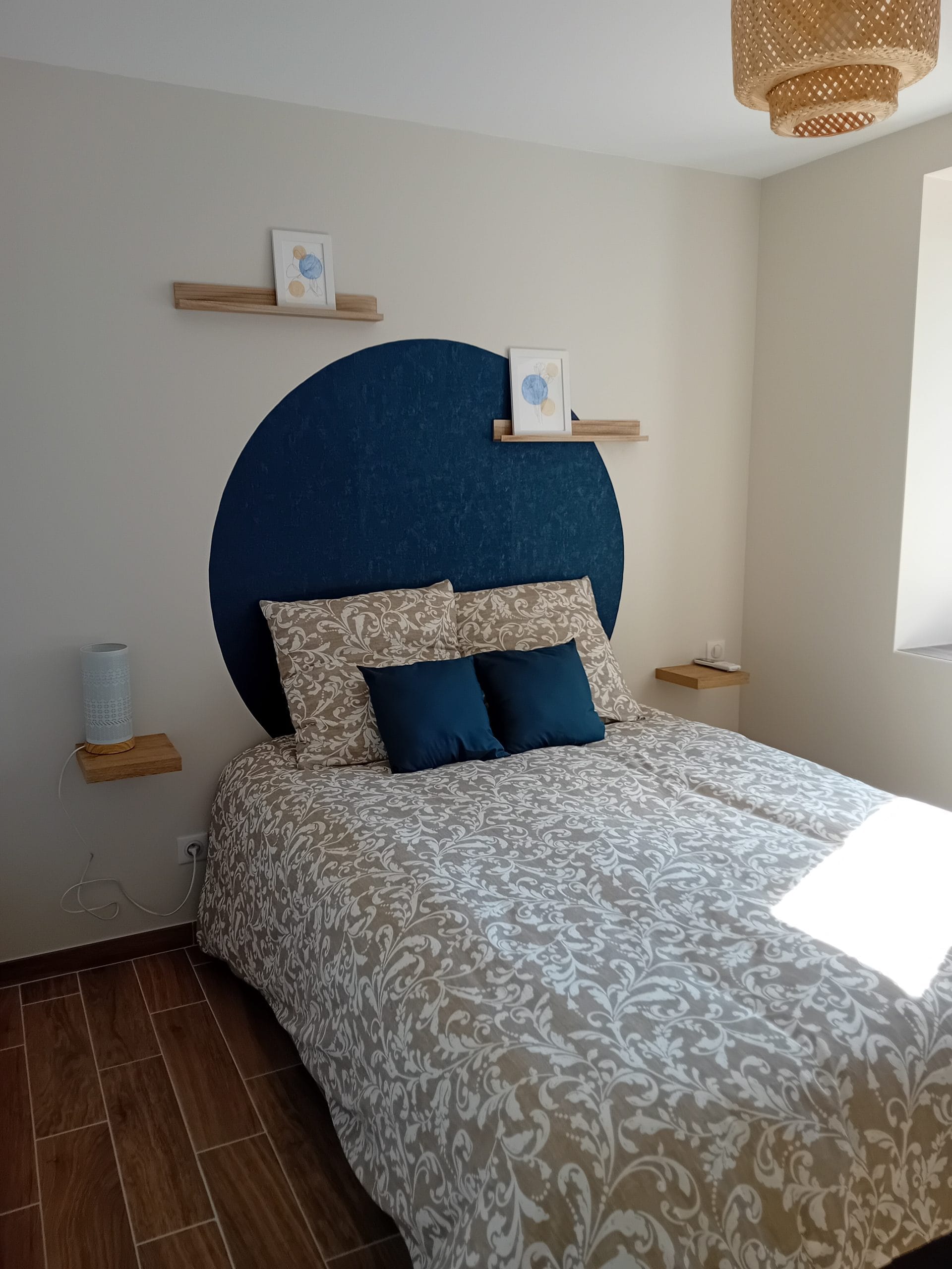 chambre avec lit double 140 x 200. murs beiges et rond bleu en guise de tête de lit avec lampe de chevet blanche. petits cadres représentant des fleurs bleues posés sur petites étagères