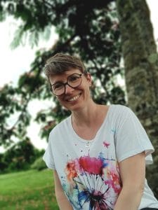 femme souriante portant des lunettes avec tee-shirt blanc et rose avec arbres et pelouses en arrière plan