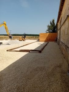 Poutres-en-bois-délimitant-un-futur-terrain-de-pétanque, avec une pelleteuse qui prend des petites pierres et rouleau compresseur derrière