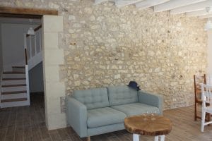 Canapé gris vert d'eau avec table basse en bois devant un mur de pierres apparentes. en arrière plan on voit un escalier bois et blanc