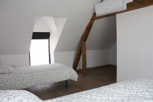 lit deux personnes au premier plan et lit une personne en arrière avec des draps fleuris noirs et blancs. sol en parquet couleur bois, poutre apparentes avec bloc de climatisation et fenêtre de lucarne grise.