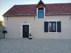 maison aux murs beiges avec porte d'entrée et fenêtres grises, cour en pierres blanches et pots de fleurs