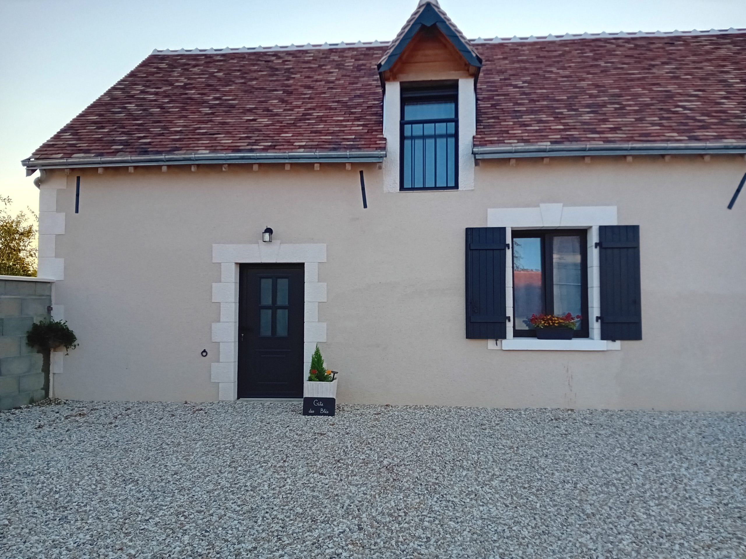 maison aux murs beiges avec porte d'entrée et fenêtres grises, cour en pierres blanches et pots de fleurs