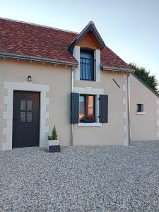 maison aux murs beiges avec porte d'entrée et fenêtres grises, cour en pierres blanches et pots de fleurs