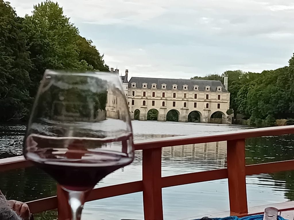 Verre de vin au premier plan avec la rambarde d'un bateau , en arrière plan on voit le château de Chenonceau avec ses arches sur le Cher