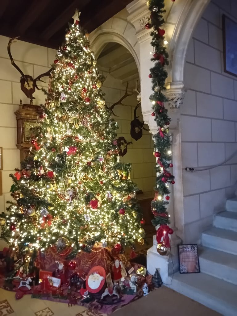 Grand sapin de noël décoré avec des boules rouges et des guirlandes lumineuses situé en bas d'un escalier en pierre.